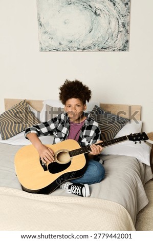 A boy sits on his bed, strumming his guitar, creating music in his personal space.