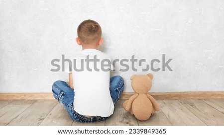 a boy sits with a teddy bear near a light wall in an empty room, back view
