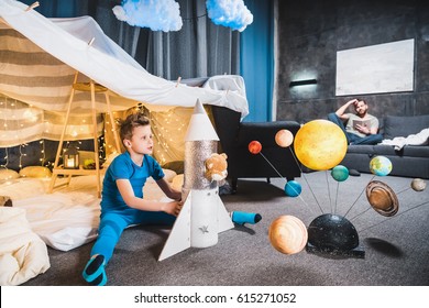 boy sitting in blanket fort with toy rocket and solar system model, father sitting on sofa behind  - Powered by Shutterstock