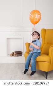 The Boy Is Sitting By The Fireplace In The Yellow Chair With Glasses Like Harry Potter, With One Balloon Celebrating Birthday.