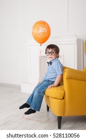 The Boy Is Sitting By The Fireplace In The Yellow Chair With Glasses Like Harry Potter, With One Orange Balloon Celebrating Birthday.