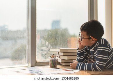 The Boy Sitting By The Window Is Using His Thoughts And Gaze To Look Outside, Doing His Homework Intently, Writing And Painting On The Table In An Art Class. 