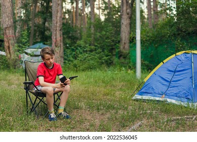 a boy sitting in a folding chair and holding a smartphone against the background of summer nature. the photo shows that even in nature and on vacation, children do not part with gadgets - Shutterstock ID 2022536045