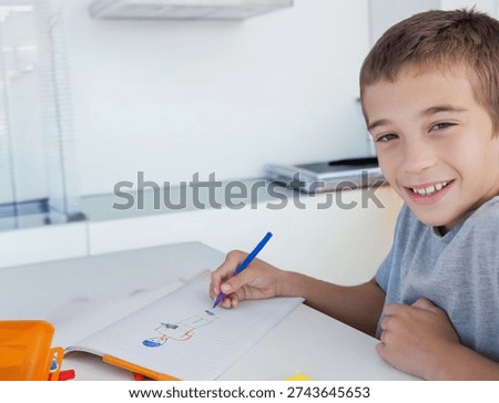 Boy sitting in kitchen drawing in notebook with blue pen and orange pencil case, copy space. Kid, drawing, sketchbook, stationery, colorful, modern, learning
