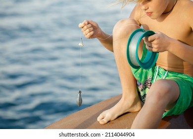 The boy is sitting on a boat with a reel of fishing line in his hands, a bait and a hook are ready on the fishing line, he is preparing to fish, there is no fishing rod