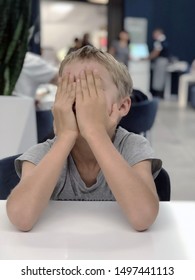 Boy Sitting On Chair Closing Face With Hands. Unhappy, Upset Kid. Bad Kids Behaviour In Public Places 