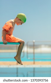 Boy Sitting On A Diving Platform Looking Down Into The Swimming Pool. Boy In Swim Gear Sitting On Spring Board.