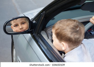 A boy, sitting on a driver place in an open top car, looks into the rear view mirror