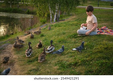 Boy sitting on the grass feeding ducks and pigeons near a pond. Outdoor nature scene. Wildlife and recreation concept. - Powered by Shutterstock