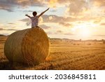 Boy sitting on a hay bale with arms raised in summer watching the sunset concept for worship, praise, religion or carefree childhood