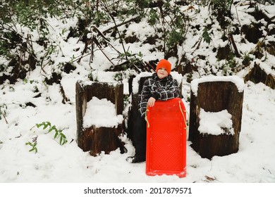 Boy Sitting On Log Seat In The Snow Holding Red Toboggan