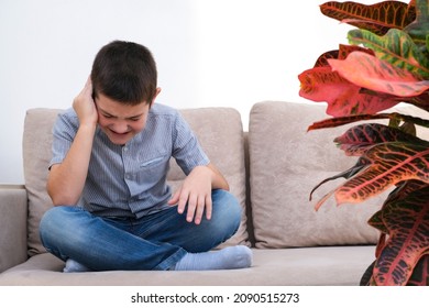 The boy is sitting on the sofa and holding his ear. the boy has an earache, he has otitis media.