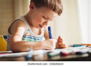 The boy is sitting at the table and diligently draws something with felt-tip pens. The child is trying so hard that he stuck out his tongue.
