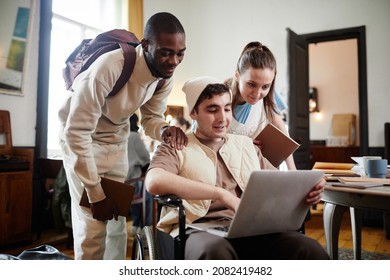 Boy sitting in wheelchair searching for information in laptop together with his two friends while they working in the library