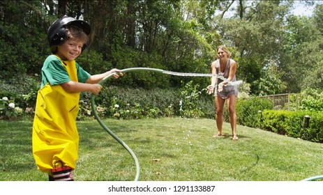 A Boy Smiles At Camera While Spraying His Mom. He Is Playing Dress Up As A Fireman