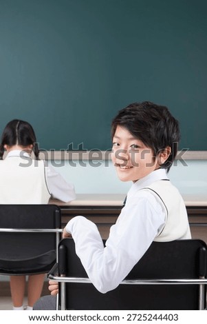 Boy smiles while sitting in classroom during lesson as classmate writes at desk
