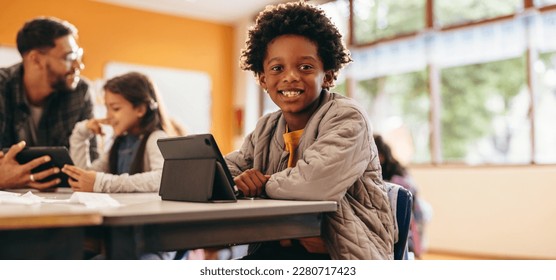 Boy smiling at the camera while sitting with a digital tablet in a classroom. Primary school kid reads an ebook at school. Young child developing his reading skills using modern technology. - Powered by Shutterstock