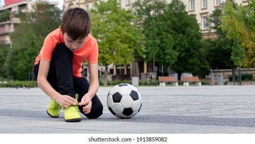 Boy soccer player tying shoelaces and soccer ball near him in town square, blurred background of cityscape in background. Sports games and leisure outside for children concept. Selective focus - Powered by Shutterstock