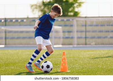 Boy In Soccer Uniform Practice With Ball. Child Kicking Ball On Grass Stadium. Young Athlete Improving Football Dribbling Skills On Training Field. Kid Soccer Player Dribbling Through Cones