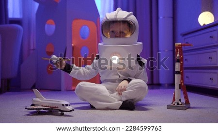 Boy in spaceman costume and helmet on his head plays with toy space satellite sitting on floor of house in evening in background of large rocket. Boy dreams of flying into space. Cosmonautics Day.