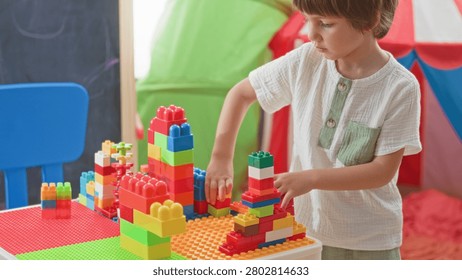 Boy stacking colorful plastic blocks with hands on a play table in a building playroom with tent and chair visible; concentration creativity.