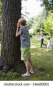 Boy standing by tree with eyes covered as children running in the background