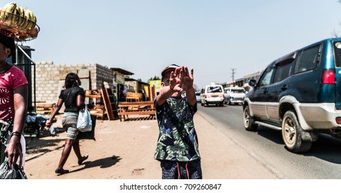 A Boy Is Standing Still With His Hands Blocking His Face. People Around Him Are Rushing Past. A Street Vendor With Bananas Walks By While Cars And Minibuses Speed On The Road.