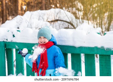 
A Boy Stands By A Green Fence In Winter. Child Holds A Snowball In One Hand. Snowballs Are Flying Into The Boy's Jacket.