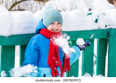 A Boy Stands By A Green Fence In Winter. Child Holds A Snowball In One Hand. Snowballs Are Flying Into The Boy's Jacket