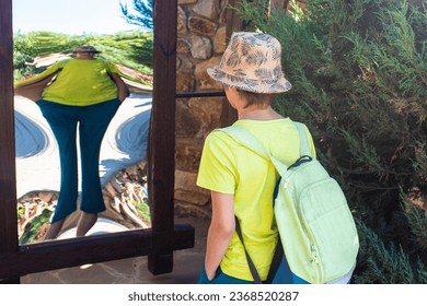 the boy stands in front of a distorting mirror. a child looks at his distorted image in a lying, false, crooked mirror in the park in summer