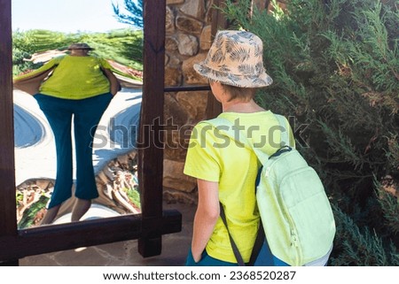 the boy stands in front of a distorting mirror. a child looks at his distorted image in a lying, false, crooked mirror in the park in summer