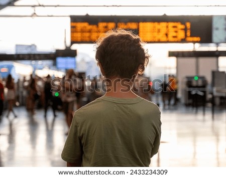 A boy stands with his back to the camera, watching the busy scene at a train station bathed in sunset light.
