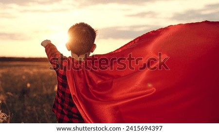 Boy stands in superhero pose with cape fluttering in wind on meadow. Calm boy with cape simulates flight of superhero in rural area. Playing favorite character and realizing of boy with cape on field