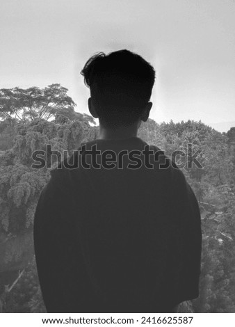 The boy stood facing the glass wall alone looking at the view outside. Boy in silhouette in room. Contemplating looking out.