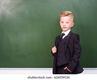 Boy in a suit standing near empty green chalkboard.