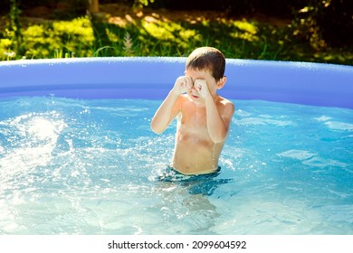 A boy swims in an inflatable pool in the garden on a sunny summer day. He rubs his eyes, from the water that has got in.