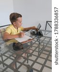 Boy talking with classmate during video call on tablet and discussing homework while sitting at table in modern light room.