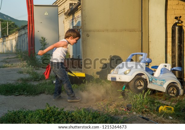 A boy in a tank, jeans and a baseball cap in the\
garage repairing the car.