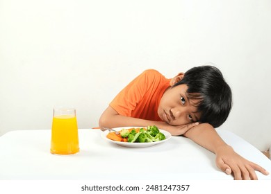 A boy with a tired expression rests his head on the table next to a plate of vegetables and a glass of orange juice.  His expression shows his dislike for the healthy meal. - Powered by Shutterstock