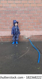 Boy Todler In Rubber Boots And Jacket Against The Background Of A Brick Wall