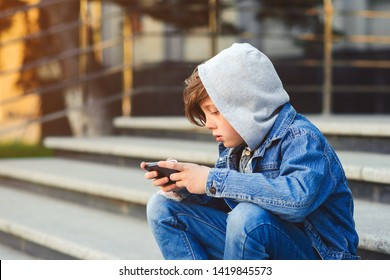 Boy use phone and plays games. Kid in the hood sitting on the stairs. Kids addicted online games and cartoons. Schoolboy plays on smartphone after school - Powered by Shutterstock