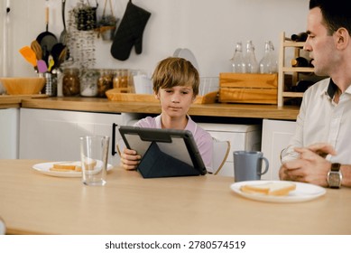 A boy uses a tablet at the kitchen table while an adult watches nearby, with plates of toast and cups on the table in a modern home setting.