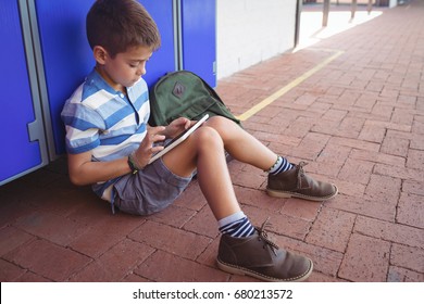Boy using digital tablet while sitting by locker in corridor at school