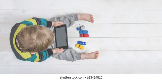 Boy using ipad / laptop while playing with toy cars on a carpet at home. Tablet pc hero header image. Boy using digital tablet while lying on wooden floor.