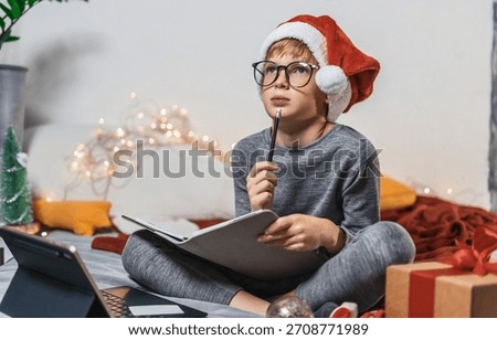 Boy using tablet for online schoolwork during Christmas holidays, sitting in cozy festive bedroom decorated with lights, gifts, and ornaments. Child writing letter to santa, christmas spirit