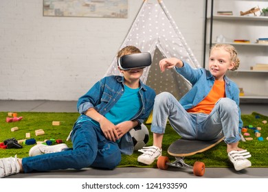 boy using virtual reality headset and his sister pointing by finger while sitting on skateboard near wigwam at home