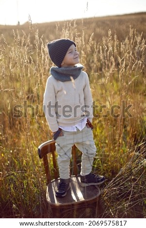 boy in warm clothes stand on chair along a path on a field with dried grass in autumn