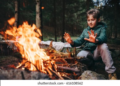 Boy warms his hands near campfire in forest
