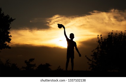 Boy Waving His Hat To The Setting Sun. Silhouette Of A Child At Sunset