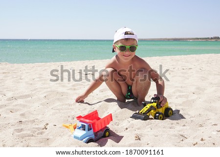 boy wearing a baseball cap and sunglasses plays with cars on a sandy beach, against a beautiful azure sea and sky on a Sunny day.